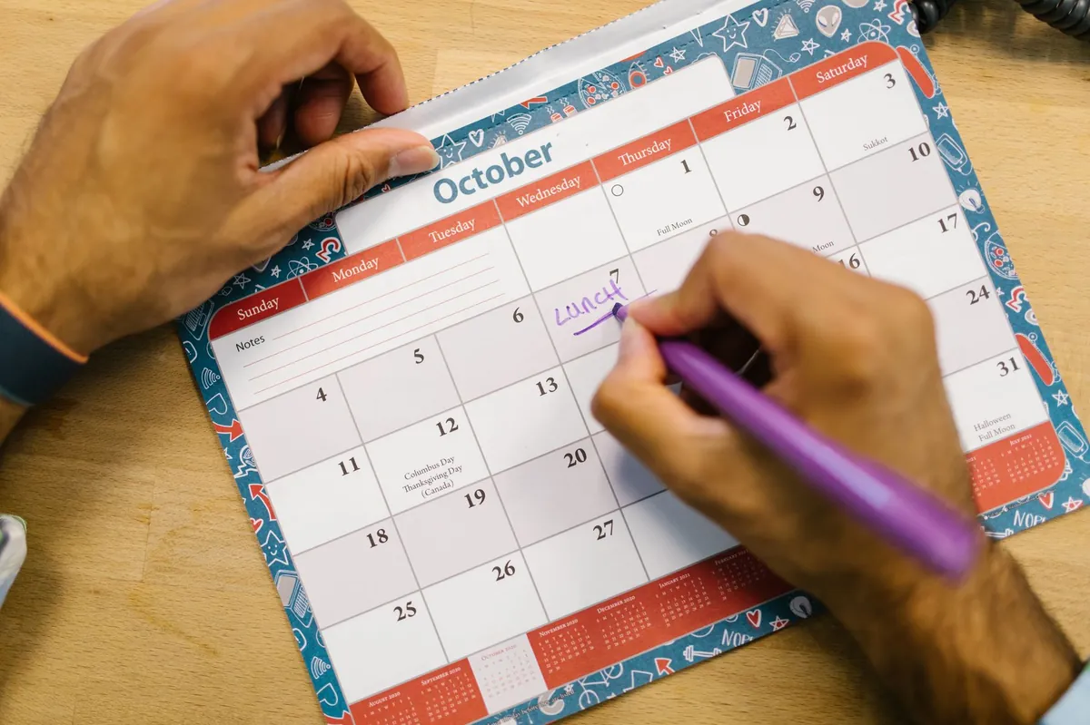 A close-up of a hand using a purple pen to write a lunch reminder on an October calendar page.